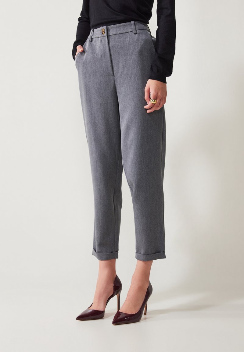 Femme portant un pantalon gris taille haute sur mesure avec des ourlets retournés, associé à un haut noir et des chaussures à talons hauts en cuir verni rouge foncé.