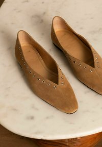 Tan suède flats met een spitse neus, voorzien van metalen oogjes langs de zijkant en een gladde binnenkant, rustend op een marmeren oppervlak.