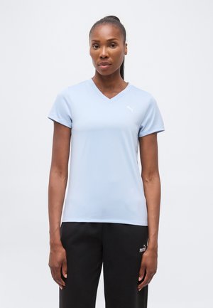 Sötét bőrű, hátrakötött hajjal rendelkező nő, aki világoskék, rövid ujjú, V-nyakú Puma pólót és fekete Puma nadrágot visel, fehér háttér előtt áll.