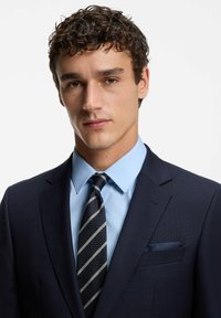 Giovane uomo con capelli ricci e scuri, che indossa un abito blu navy, una camicia azzurro chiaro e una cravatta rigata scura, rivolto verso lo spettatore su uno sfondo semplice.