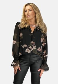 Czarna bluzka w kwiaty z długimi przezroczystymi rękawami, falbanami na mankietach i dekoltem w serek. Posiada jasny wzór kwiatowy w odcieniach różu i zieleni.