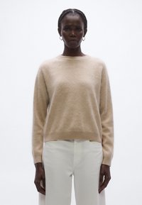 Beżowy sweter z kaszmiru z okrągłym dekoltem, długimi rękawami i prążkowanym obszyciem. W zestawieniu z białymi spodniami, na neutralnym tle.