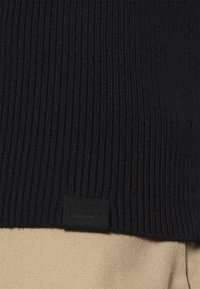Black ribbed sweater made of textured knit fabric, with a small black label featuring "PIER ONE" at the bottom. Background shows beige fabric.