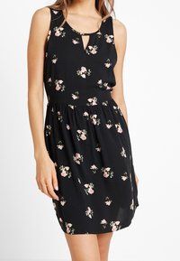 Ärm Lös svart klänning med samlad midja och små rosa blommor, bärs av en stående person från halsen till mitten av låret.