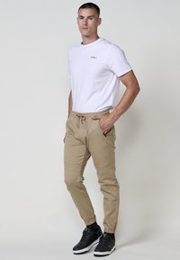 Bărbatul stă cu mâinile în buzunare, îmbrăcat cu un tricou alb, pantaloni jogger bej, pantofi sport negri și șosete închise la culoare, pe un fundal simplu.