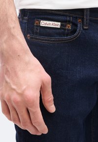 Κοντινό πλάνο ενός χεριού δίπλα στην μπροστινή τσέπη σκούρου μπλε τζιν με εμφανή ετικέτα Calvin Klein.