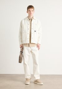 White denim jacket with multiple pockets, beige plaid shirt, white trousers, and beige sneakers. Accessory: small beige shoulder bag.