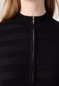 Gros plan d'une personne portant un pull noir à fermeture éclair avec des rayures horizontales discrètes et un col montant côtelé.
