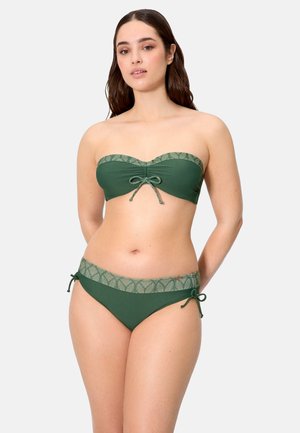 Mujer con bikini bandeau verde con ribete estampado y lazos anudados en el centro superior y en los lados inferiores, de pie frente a un fondo liso.