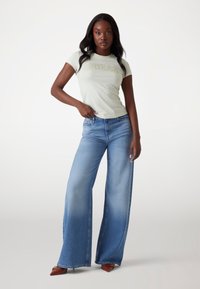 Lys grøn tætsiddende t-shirt med "GUESS" logo, parret med vidde ben blå denim jeans med en falmet vask og spidse brune sko.