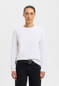 Weißes langärmeliges Baumwollshirt mit rundem Ausschnitt, kombiniert mit dunklen Jeans aus Denim und einem schwarzen Gürtel mit silberner Schnalle.