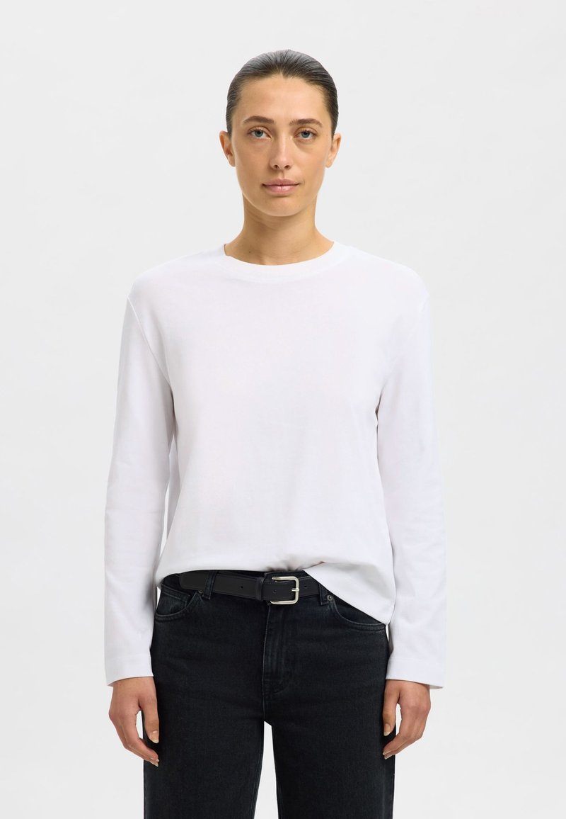 Weißes langärmeliges Baumwollshirt mit rundem Ausschnitt, kombiniert mit dunklen Jeans aus Denim und einem schwarzen Gürtel mit silberner Schnalle.