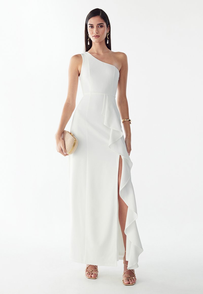 Woman wearing a white one-shoulder gown with a ruffled thigh-high slit, holding a beige clutch, and wearing gold jewelry and strappy heels.