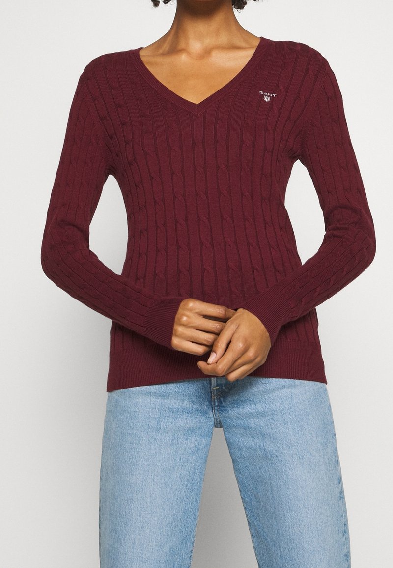 Femme portant un pull en maille torsadée bordeaux à col en V, associé à un jean bleu clair, les mains jointes devant elle.