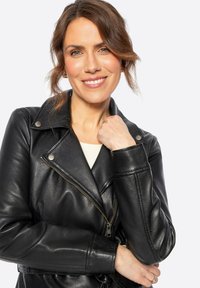Chaqueta de motocicleta de cuero negro con herrajes plateados, cierre de cremallera al frente, solapas anchas y un corte estructurado. Textura suave con un brillo sutil.