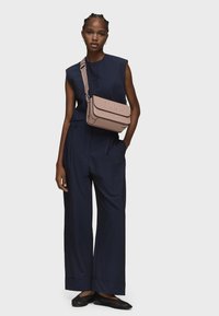 Borsa a tracolla in morbida pelle rosa pallido con logo "TOUS" in rilievo, arricchita da una tracolla removibile. Il modello indossa una maglia over-size blu navy e pantaloni larghi.