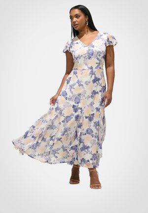 Vrouw die een vloeiende, enkellange jurk met bloemenpracht draagt, met korte, vliegende mouwen en sandalen met hoge hakken en bandjes, staand tegen een effen achtergrond.