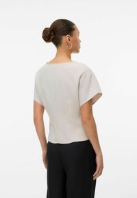 Lys beige topp med korte, vide ermer og rund hals. Har en jevn tekstur og en skreddersydd passform ved livet, kombinert med svarte bukser.