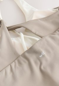 Close-up of beige and white fabric with overlapping straps and the word "self." printed in white on beige material.