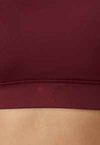 Bordo sportski grudnjak s glatkom elastičnom završnom obradom, s logotipom Puma u malo tamnijoj nijansi. Jednostavan dizajn i usko pristajanje.
