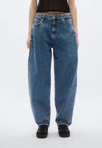 Laisvai priglundančios mėlynos spalvos džinsinės kelnės su aukštu liemeniu, turinčios priekines kišenes ir sagų užsegimą, derinamos su juodais sportbačiais kontrastui.