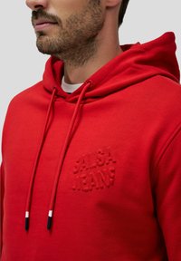 Sudadera roja hecha de tejido suave con un bolsillo tipo canguro; presenta un logo texturizado de 'Salsa Jeans' en el pecho y cordones con puntas de color.