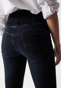 Žena v tmavě modrých úzkých vysokých džínech a bílém tričku s dlouhým rukávem s vyhrnutými rukávy, zobrazená ze zadu.