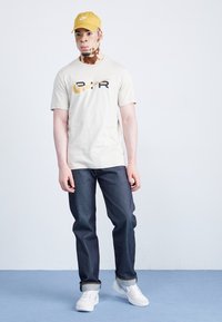 Beige tričko s černým a žlutým logem, tmavé džíny a bílé tenisky. Model má na sobě žlutou kšiltovku a stojí na modré podlaze.