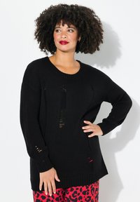 Schwarzer Strickpullover mit lockerer Passform, der Schnittdetails auf der Vorderseite und gerippten Bündchen aufweist. Kombiniert mit roten Leopardmuster-Hosen.