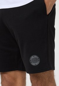 Juodi medvilniniai šortai, tinka prie kūno. Kairėje kojoje yra apvalus juodas pleistras su baltu tekstu ir pasaulio grafika.