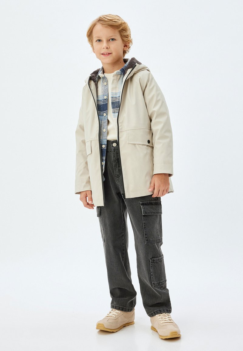 Beige vattentät jacka med huva, framdragkedja och fickor, bärs över en blå rutiga skjorta, ihop med mörka cargo-byxor och beige sneakers.