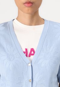 lichtblauwe gebreide cardigan met V-hals, bloemenpatroon en gouden knopen, gedragen over een crèmekleurig shirt met roze kraalletters.