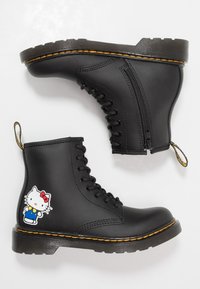 Černé kožené šněrovací boty s bočním zipem, žluté stehování na podrážkách a nášivka postavy Hello Kitty na vnější straně jedné boty.