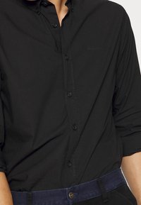 Camicia nera con bottoni, realizzata in tessuto morbido, caratterizzata da un colletto a punta e maniche lunghe, con una delicata ricamo del logo vicino alla tasca sul petto.