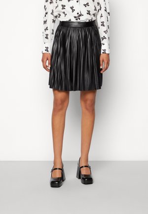 Femme portant une blouse blanche avec des imprimés de nœuds noirs, une jupe plissée en cuir noire et des escarpins Mary Jane à talon bloc noirs, debout sur un fond blanc.