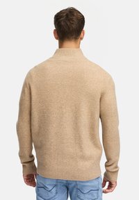 Beiger Strickpullover mit gerippten Bündchen und Saum. Weicher Stoff, hoher Kragen und lässige Passform, getragen mit hellblauen Jeans.