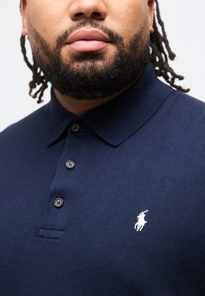 Άνδρας με γενειάδα που φοράει μπλε ναυτικό πόλο με τρία κουμπιά και λευκό κέντημα λογότυπου με άλογο και ιππέα.