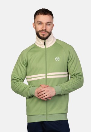 Groene ritssluitingjack met crèmekleurige hoge kraag en twee horizontale witte strepen. Glad materiaal met geribde manchetten en een logo op de borst.