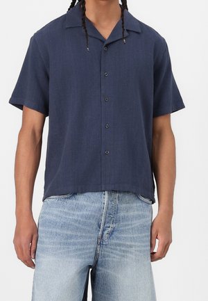 Mand iført en navy kortærmet skjorte med knapper og lyseblå denimjeans mod en ensfarvet baggrund.