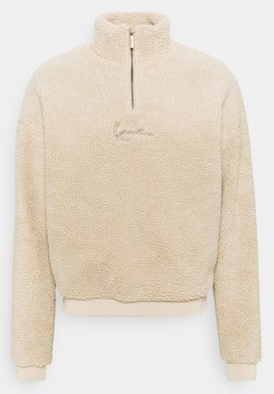 Mann trägt beigen Fleecepullover, weißes Hemd darunter, braune Velourhosen, weiße Socken und weiße Turnschuhe vor grauem Hintergrund.