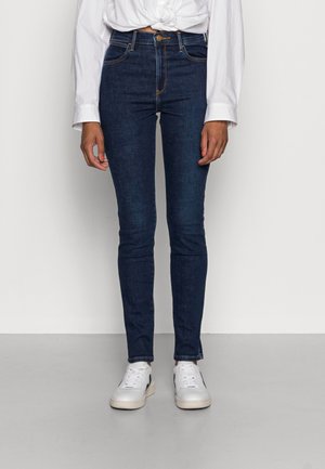 Jeans ajustados de mezclilla azul oscuro, con una cintura alta, diseño de cinco bolsillos y costuras en contraste, combinados con zapatillas blancas.