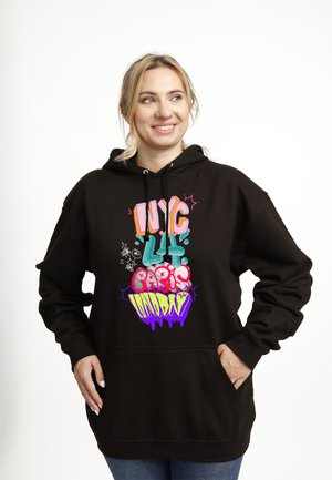 Lächelnde Frau mit blonden Haaren trägt schwarzen Hoodie mit buntem Graffiti-Text, der NYC, LA, Paris und London auflistet, und steht vor weißem Hintergrund.