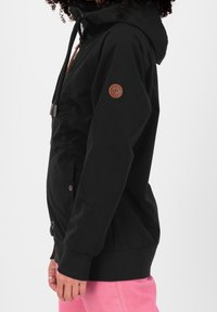 Persona che indossa una giacca nera con cappuccio e una toppa rotonda marrone sulla manica, con pantaloni rosa, in posizione di profilo su uno sfondo bianco.