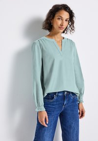 Femme aux cheveux courts et bouclés, portant un haut à manches longues vert clair et un jean bleu, se tenant contre un mur blanc avec une douce ombre.
