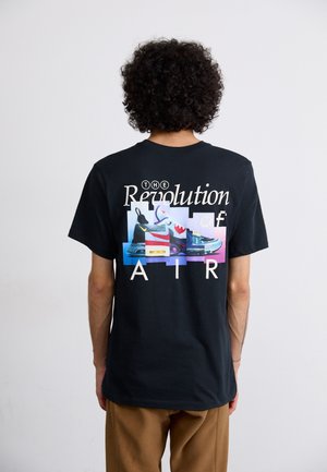 Osoba z kręconymi włosami nosi czarną koszulkę z kolorową kolażem sneakersów i tekstem "The Revolution of Air" na plecach.