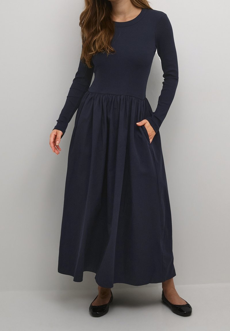 Femme portant une longue robe bleu marine à manches longues et une jupe froncée, debout avec une main dans sa poche et des ballerines noires.