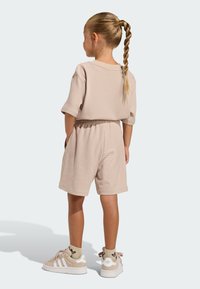 Béžový rebrovaný top s krátkym rukávom a zodpovedajúce šortky s elastickým pásom. Modelka má na nohách béžové tenisky a svetlé vzorované ponožky.