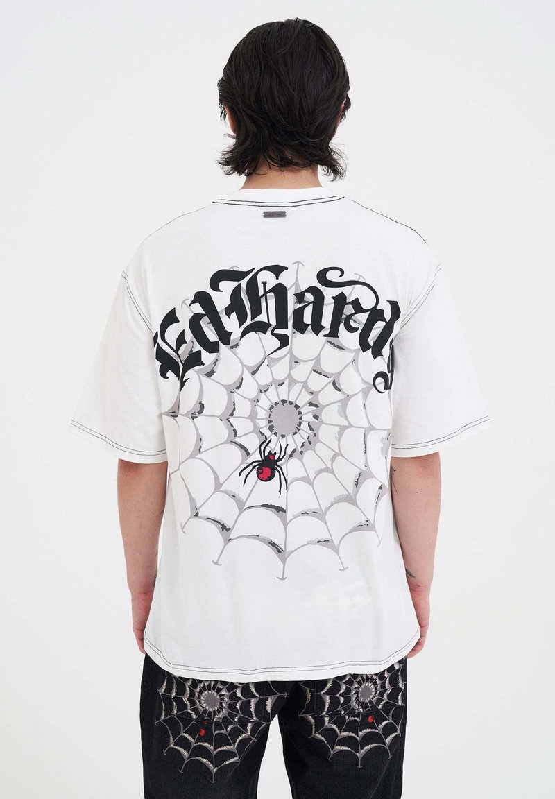 Hvid oversized t-shirt med sort gotisk tekst og et edderkoppespind-grafik på ryggen. Har en edderkop med røde detaljer i nettet.