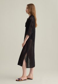 Schwarzes, gestricktes Midi-Kleid mit lockerer Passform, Dreiviertelärmeln, Schlitz an den Seiten und einem kunstvollen Lochmuster. Kombiniert mit bunten Sandalen.