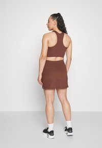 Smeđi sportski crop top s leđima u obliku racerback, uparen s usklađenom suknjom. Model nosi crne tenisice i bijele člančaste čarape.